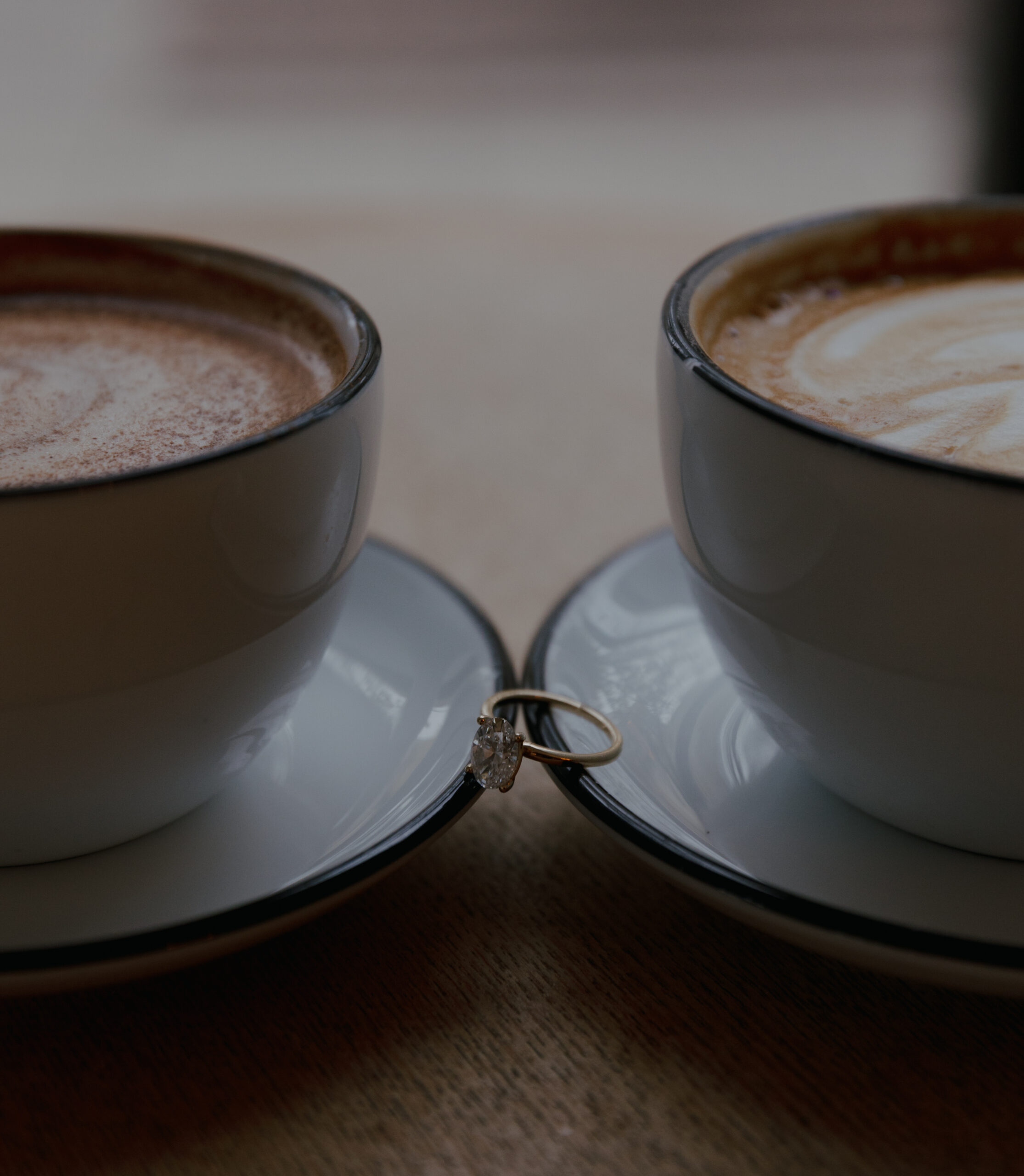 two lattes with saucers on a table with an engagement ring between the two